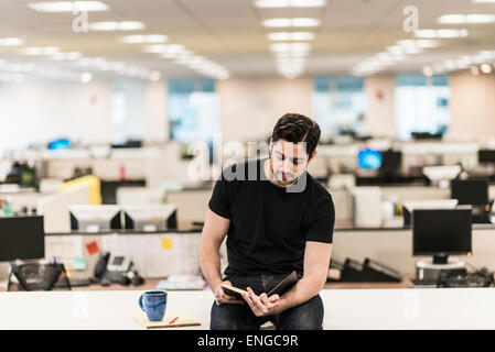 A man seated on a desk in an office looking at a book or diary. Stock Photo