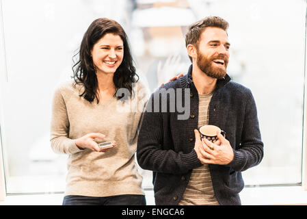 A business woman seated by a window holding a smart phone, talking to a man holding a coffee cup. Stock Photo