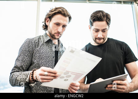 Two men standing in an office, looking at a page of printing, and referring to a digital tablet. Stock Photo