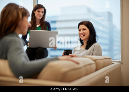 Three businesswomen by a window in an office, one holding a laptop. Stock Photo