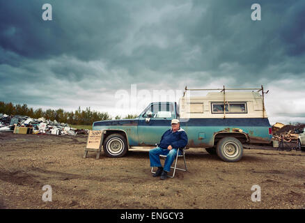 A mature man seated in a chair by his pick up truck. Piles of waste, scrap metal and wood objects. Stock Photo