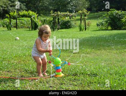 Blond curly haired girl toddler, child, playing with a toy sprinkler in the outdoor sunshine, surrounded by green lawn and trees Stock Photo