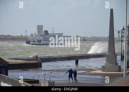 During stormy weather, a Wightlink ferry sails through the rough sea, and two people have trouble standing up due to the high winds on the promenade at Southsea, Portsmouth, Hampshire, England. Stock Photo