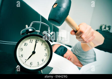 a young caucasian man in bed about to hammer his alarm clock at seven in the morning Stock Photo
