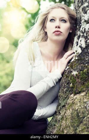 Outdoor fashion portrait of young Caucasian woman leaning against a tree Stock Photo