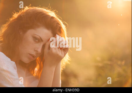 Young woman with auburn hair laying in a sunlit field at dusk Stock Photo