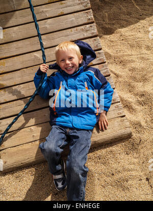 Child plays on Playground and is smiling. The boy fell on the playground . He wears his blue overcoat Stock Photo