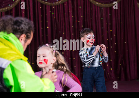 Boy Dressed as Clown Wearing Make Up and Striped Shirt Performing on Stage with Red Curtain, Girl Having Make Up Applied to Face Stock Photo