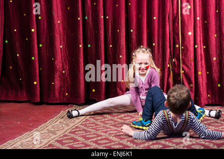 Blond Girl Wearing Clown Make Up Performing Splits on Stage with Red Curtain with Young Boy in Foreground Stock Photo