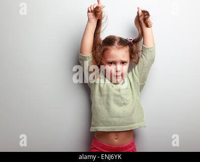 Active unhappy kid girl pulling her long hair on blue wall background with empty copy space Stock Photo