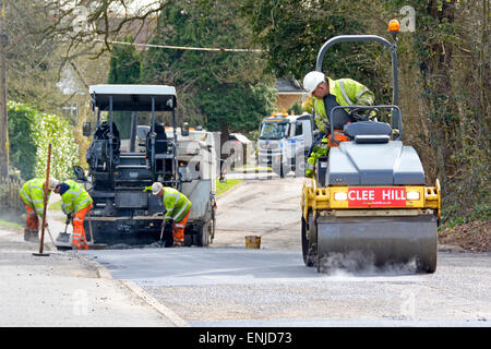 Men working at road works resurfacing & repairs using vibrating roller & tarmac spreading machine wearing high visibility jackets & hard hats Essex UK Stock Photo