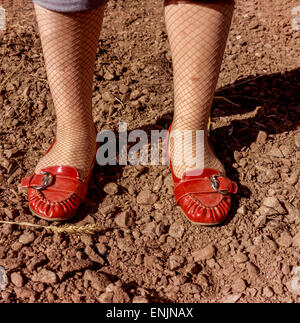 Elegant woman legs in fishnet stockings and red patent leather loafers standing on rough rocky ground, showing style contrast Stock Photo