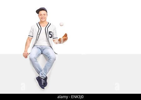 Young man playing with a baseball seated on a blank signboard and looking at the camera isolated on white background Stock Photo