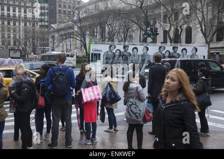 Pedestrians wait to cross 42nd St. at 5th Ave with the NY Public Library in the background along with a moving bus advertisement Stock Photo