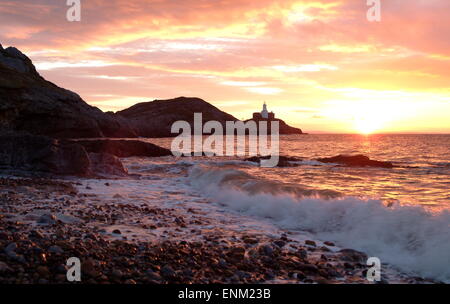 a flaming orange sunrise glints on the surf crashing onto shingle. A lighthouse and 3 small islands in the distance Stock Photo