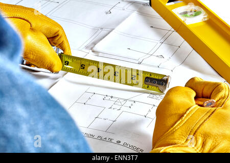 Man's hands in leather work gloves holding a tape measure and a pencil reviewing blueprints with level laying on plans Stock Photo