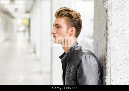 Attractive blond young man with leather jacket standing outside against pillar, looking away Stock Photo