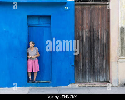A Hispanic senior citizen stands in the doorway of a deep blue wall looking sad in Santiago de Cuba. Stock Photo