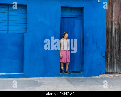 A Hispanic senior citizen stands in the doorway of a deep blue wall looking sad in Santiago de Cuba. Stock Photo