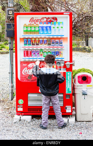 Back of Caucasian child, boy, 8-9 year old, standing in front of Japanese coca cola vending machine that towers over him, pressing button for a drink. Stock Photo