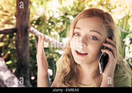 Outdoor portrait of beautiful blond Caucasian teenage girl in a park talking on a cell phone, vintage toned photo filter effect Stock Photo