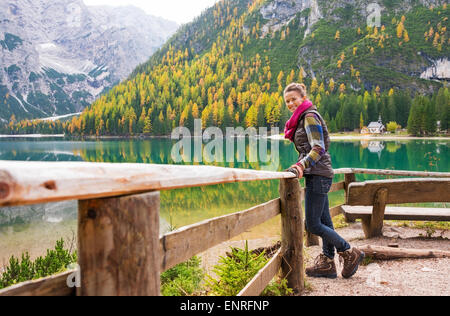 A smiling brunette hiker wearing outdoor gear stands facing the water and leaning against a wooden railing. The still water in t Stock Photo