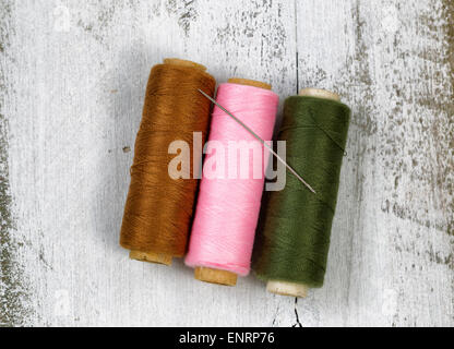 Three spools of different colors of thread, with single needle, on top of rustic white wood. Stock Photo