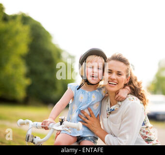 Proud moment shared between a mother and a daughter who has just learned how to ride her bicycle. The mother kneels lovingly next to her daughter, holding her gently, as her daughter throws a loving arm around her mother's neck. Both are smiling, happy, and proud. Stock Photo