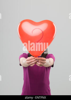 caucasian woman holding heart shaped balloon with question mark in front of her head, against grey background Stock Photo