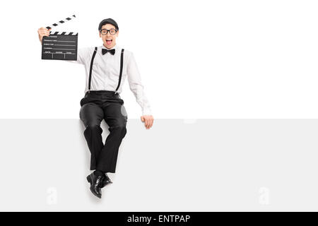 Cheerful male movie director holding a clapperboard and posing seated on a blank panel isolated on white background Stock Photo