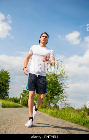 Handsome young man running and jogging on road in the country in a sunny day, wearing white shirt and baseball cap Stock Photo