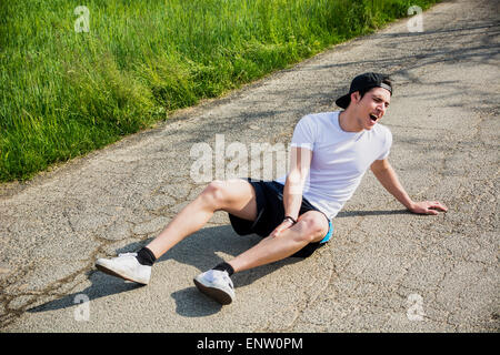 Handsome young man injured while running and jogging on road in the country in a sunny day, wearing white shirt and baseball cap Stock Photo