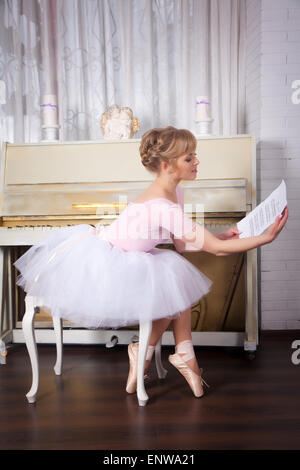Pretty ballerina with notes in her hands in pointe shoes sitting on a chair near the piano