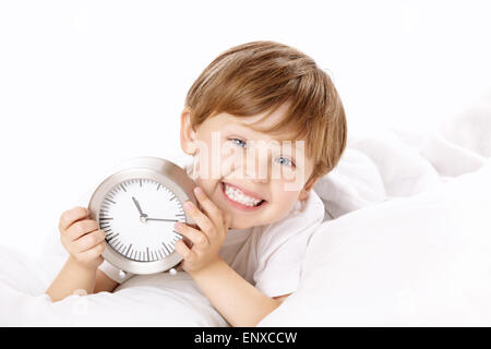 The smiling boy lies in bed with an alarm clock in hands Stock Photo
