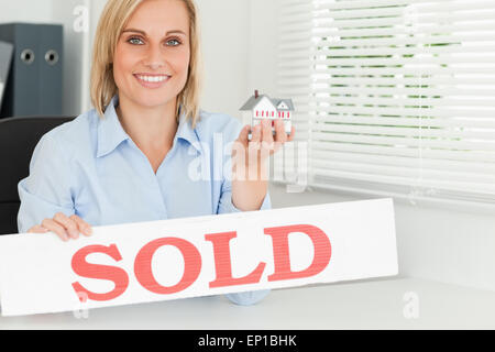 Blonde businesswoman showing miniature house and SOLD sign looking into camera Stock Photo