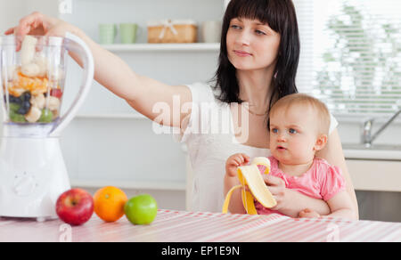 Gorgeous brunette woman putting vegetables in a mixer while holding her baby on her knees Stock Photo