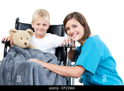 Nice female doctor carrying adorable little boy with his teddy bear in the wheelchair at the hospital Stock Photo