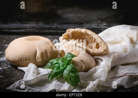 Two homemade whole and sliced pita bread stuffed with fresh basil on white textile, served with flour over dark table Stock Photo