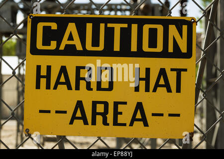 yellow and black caution hard hat area sign wired to a chain link fence Stock Photo