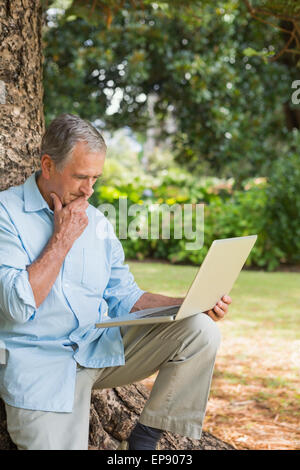 Retired man leaning against tree with a laptop Stock Photo