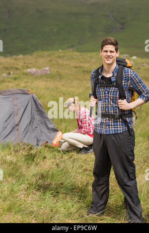Happy man carrying backpack while girlfriend is pitching tent Stock Photo