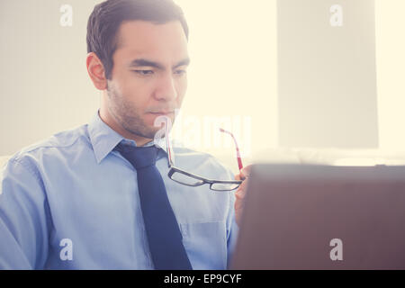 Unsmiling man biting his eyeglasses and using a laptop Stock Photo