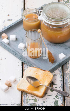 Toast with homemade caramel sauce for breakfast, served with spoon, jars and sugar cubes on gray tray over old wooden table Stock Photo