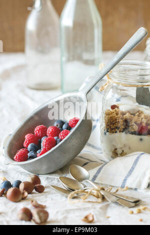 Glass jar with homemade granola and yogurt served with nuts, raspberries and blackberries in vintage colander over white textile Stock Photo