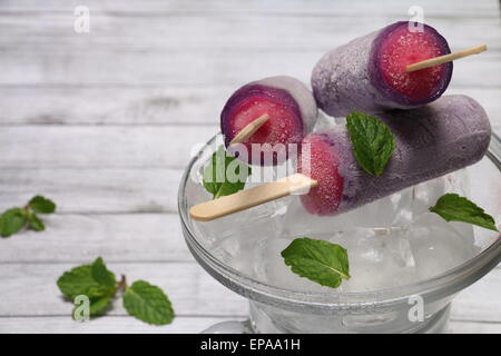 Strawberry and grape ice lollies on bed of ice cubes, garnished with mint leaves on rustic wooden board Stock Photo