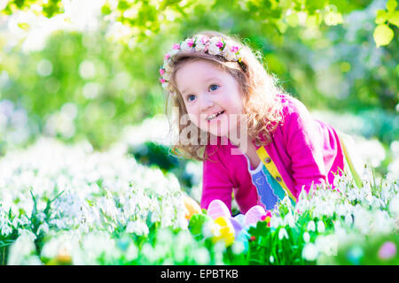 Adorable curly toddler girl in a pink summer dress playing with Easter eggs in garden with first spring flowers Stock Photo