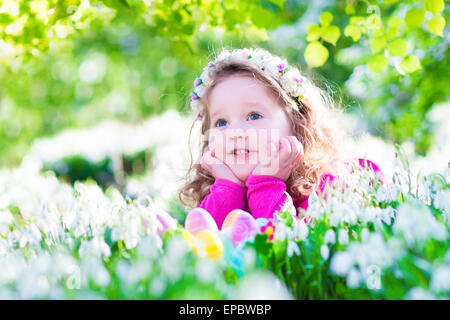 Adorable curly toddler girl in a pink summer dress playing with Easter eggs during egg hunt in garden with first spring flowers Stock Photo