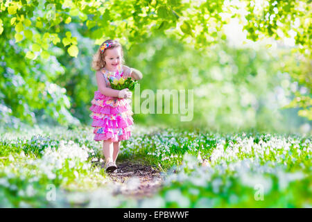 Adorable curly toddler girl in a pink summer dress playing with Easter eggs in sunny garden with first white spring flowers Stock Photo