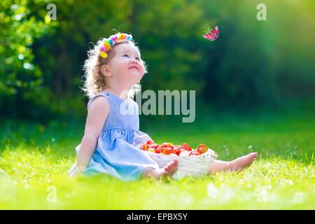Cute little toddler girl with curly hair wearing a blue summer dress having fun in the garden eating healthy fresh strawberry Stock Photo