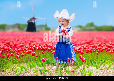 Adorable toddler girl wearing Dutch traditional national costume dress playing in field of blooming tulips next to windmill Stock Photo